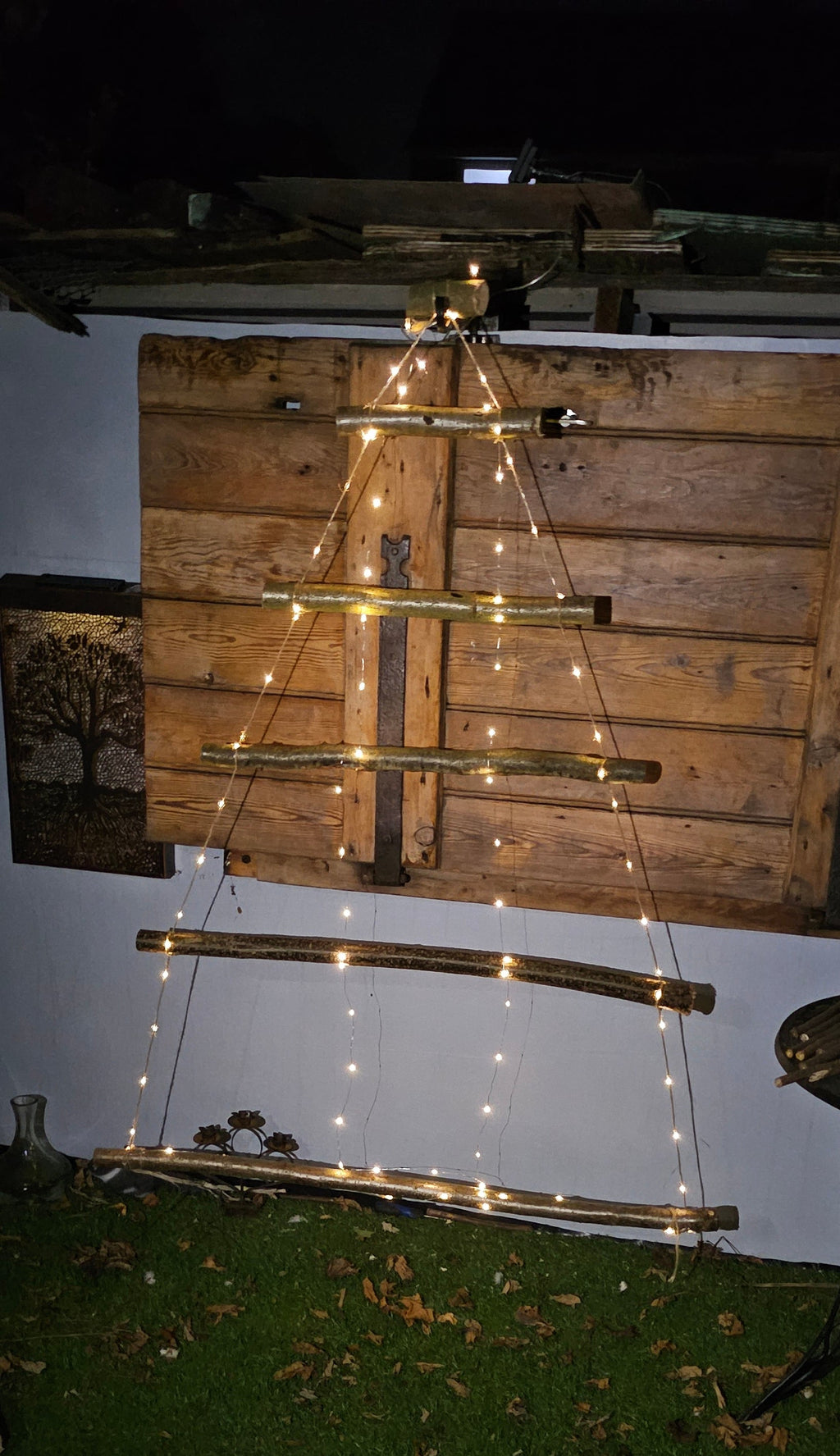 A wooden ladder-style string light decoration set up outdoors against a white wall and wood paneling, with warm glowing lights along the rungs on a dark night scene.