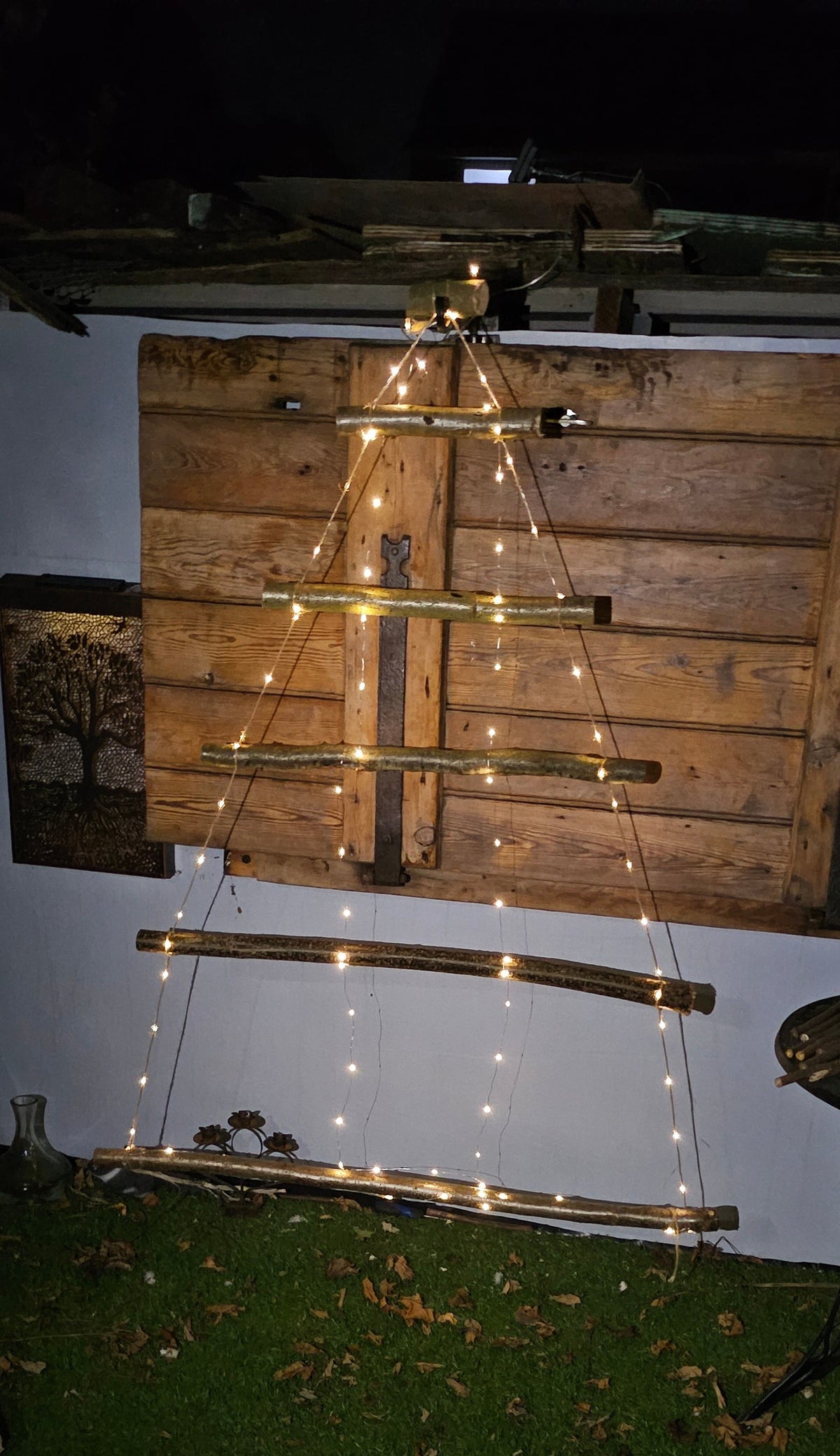 A wooden ladder-style string light decoration set up outdoors against a white wall and wood paneling, with warm glowing lights along the rungs on a dark night scene.