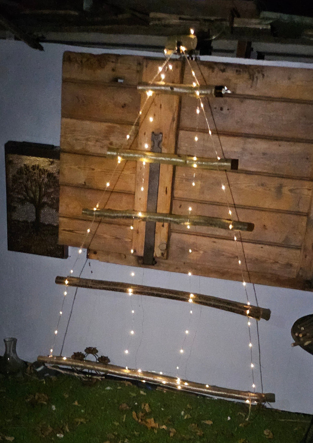 A rustic wooden ladder-style Christmas tree decoration with warm white string lights, set against a wooden pallet backdrop on an outdoor grassy lawn at night.