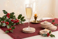 A glass of bubbly champagne on a wooden coaster, set on a red runner with holly leaves and red berries, alongside a lit candle and stacked birch wood slices.